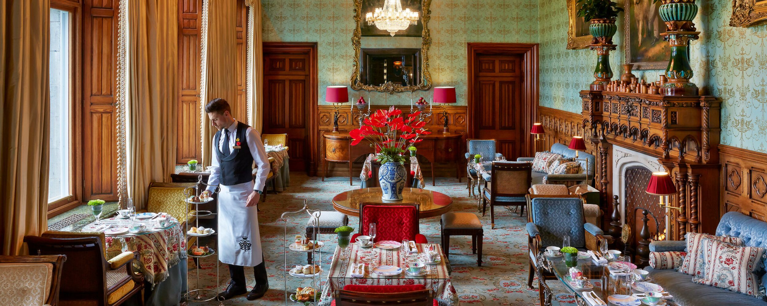 An elegant dining room featuring ornate decor, plush chairs, and a large table set for afternoon tea. A waiter in formal attire attends to the table, which is adorned with small plates and floral arrangements. Grand windows allow natural light to fill the space, highlighting intricate wallpaper and decorative chandelier.