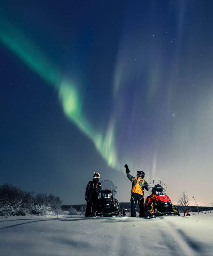 Two people on snowmobiles looking up to the green light of the northern lights