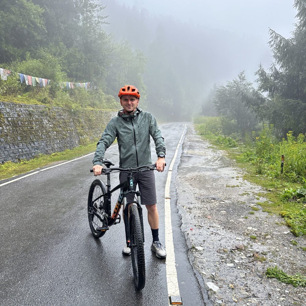 A person stands next to a mountain bike on a misty, quiet road surrounded by greenery. They are wearing a gray jacket, orange helmet, and shorts. Trees and prayer flags are visible in the background.