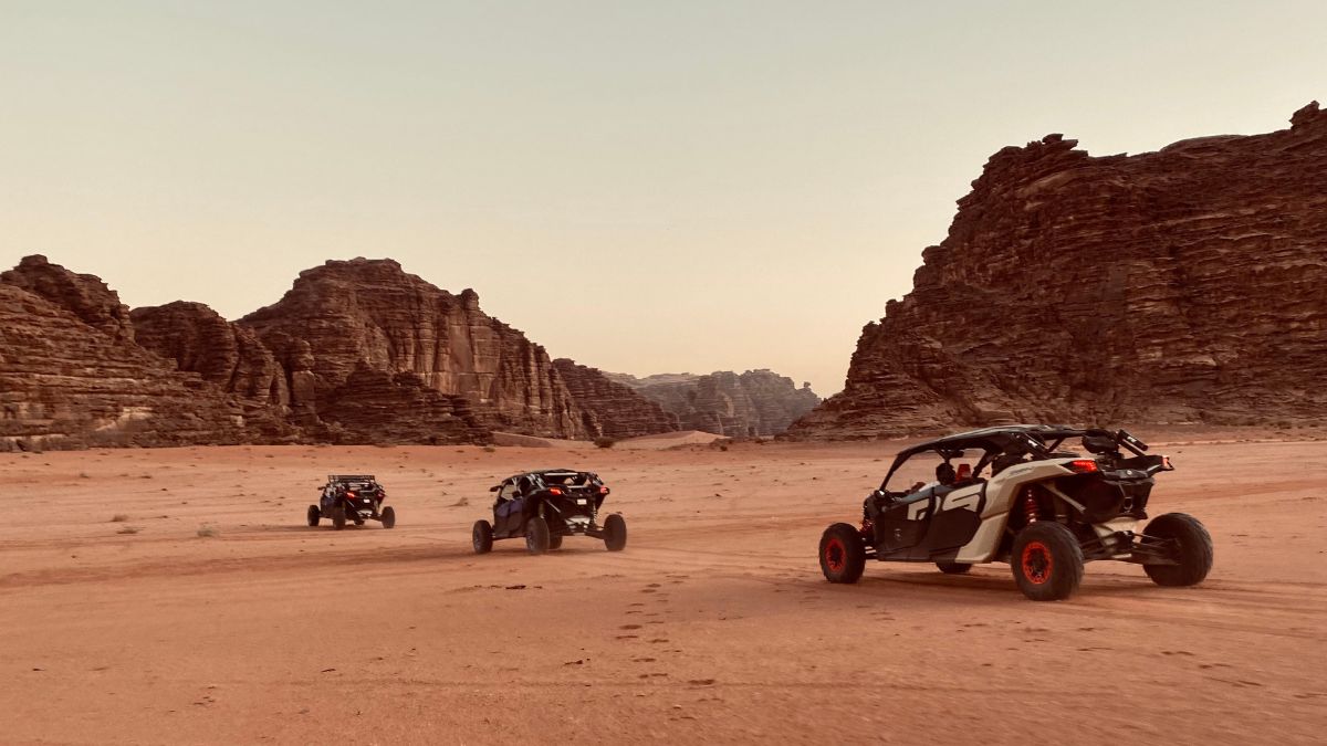 Three off-road vehicles navigate through a vast desert landscape, surrounded by towering rock formations and rugged mountains during dusk. The sandy terrain shows tire tracks, highlighting the vehicles' adventurous path.