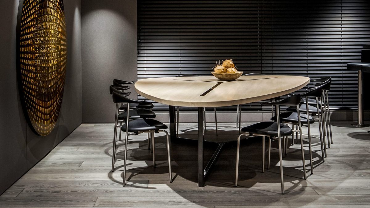 A contemporary dining area featuring a uniquely shaped wooden table with a central bowl of fruit. Surrounding the table are black chairs, and the walls have a textured design with horizontal blinds and abstract artwork.