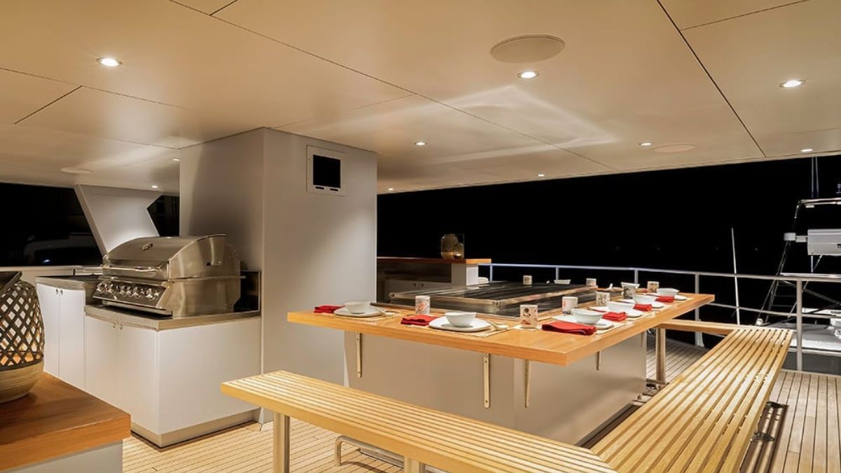 A modern outdoor kitchen on a yacht, featuring a grill, countertop, and dining tables set with plates. Soft lighting illuminates the space against a dark background, creating a cozy ambiance.