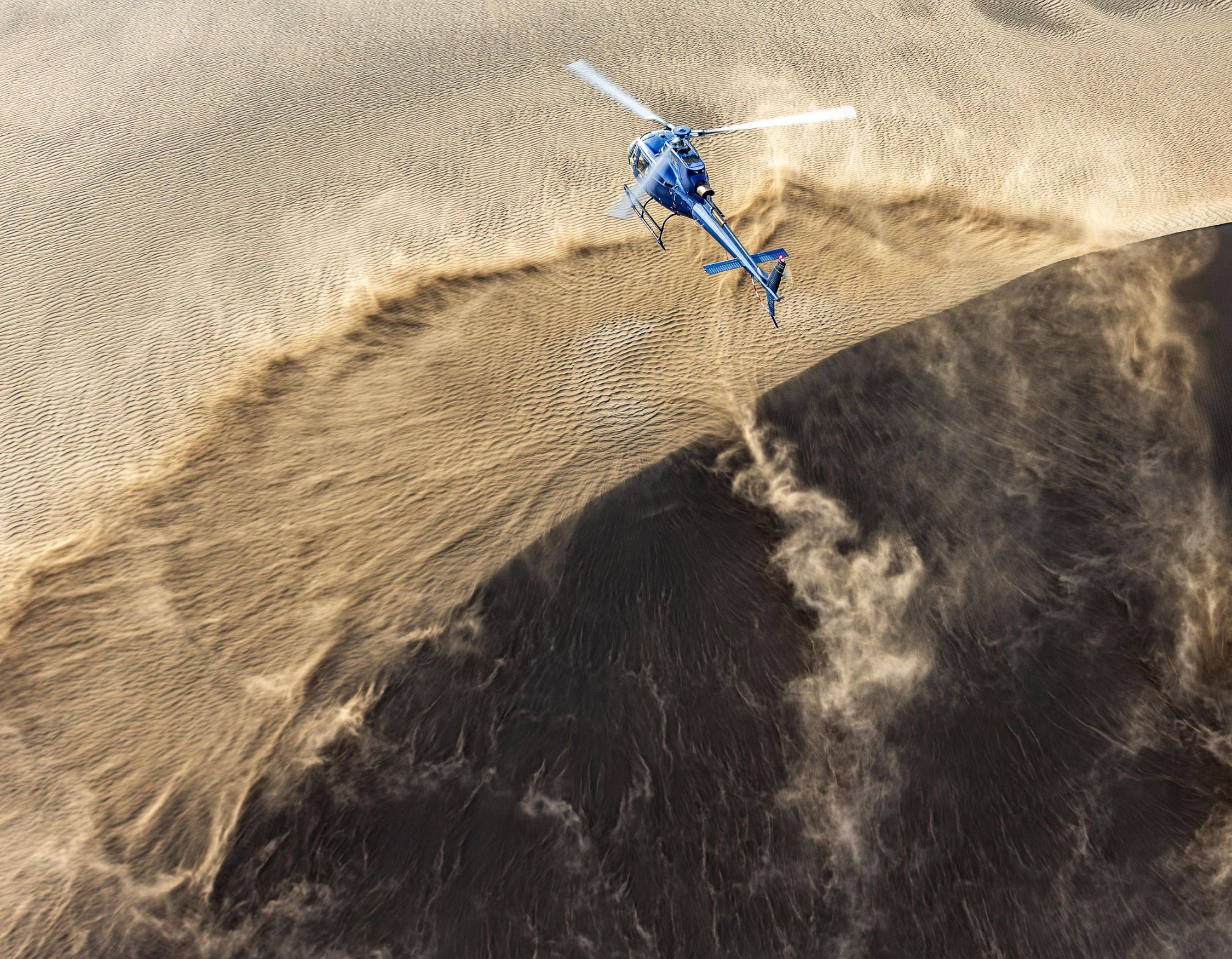 A blue helicopter flies low over a sandy landscape, creating a swirl of dust and debris in its wake. The contrasting colors of sand and shadows highlight the curvature of the terrain.