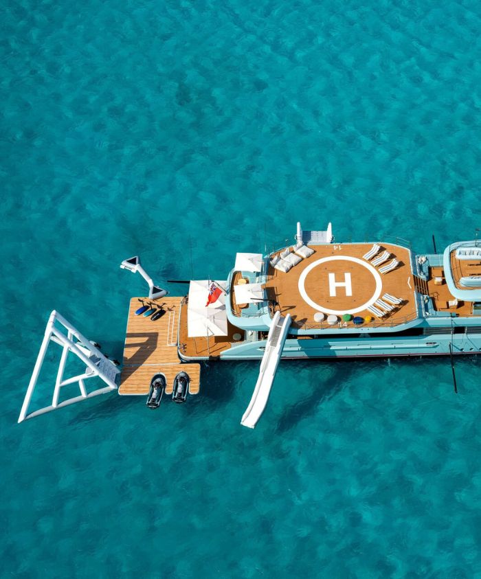 Blue luxury boat with a sun deck and sun loungers on a heli pad. Also an adventure park off the back of the boat in clear blue waters.