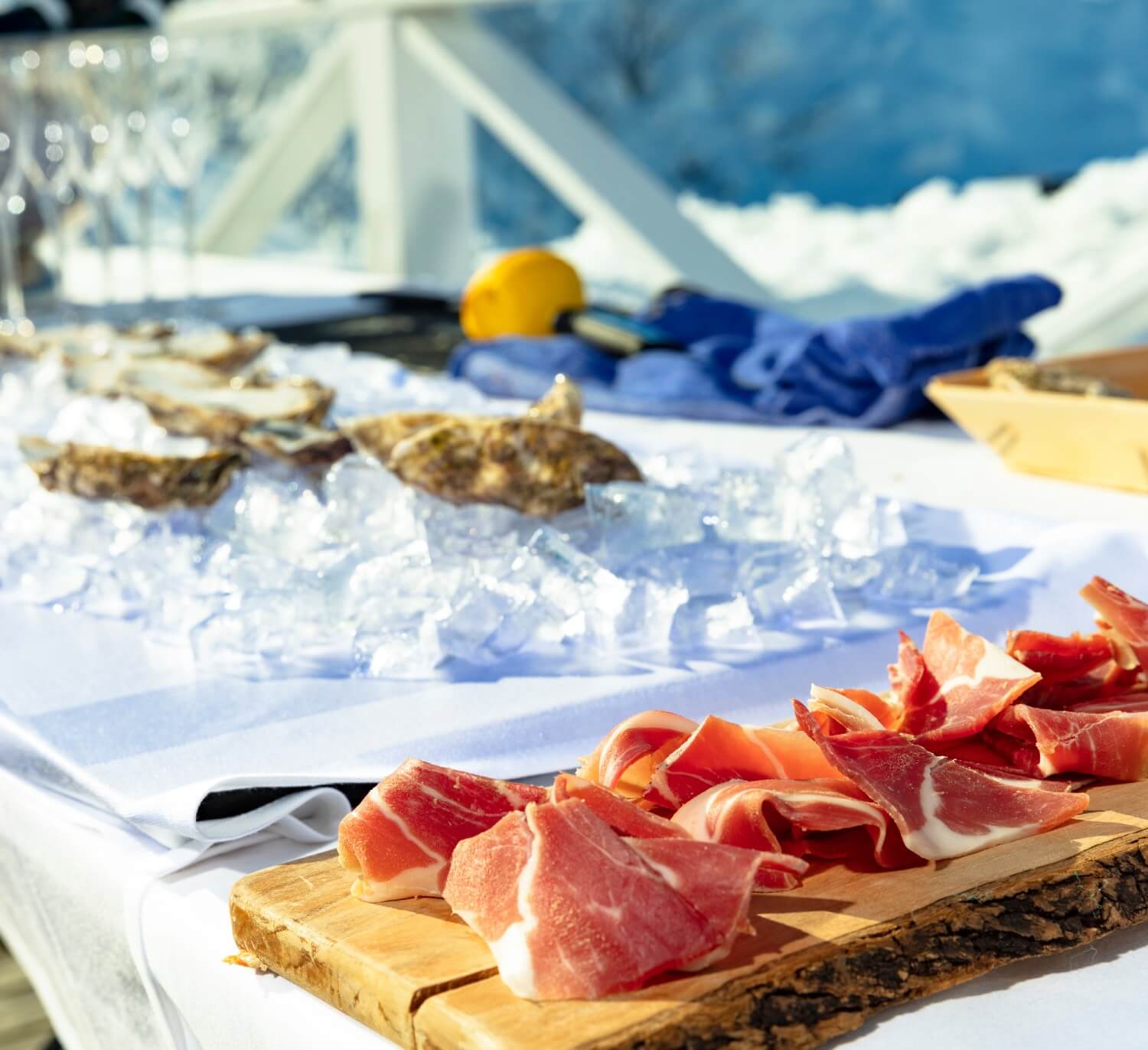 A beautifully arranged food display featuring thinly sliced cured meats on a wooden board, with oysters and seafood on ice in the background. The setting has a bright, sunny ambiance, complemented by snow in the distance.