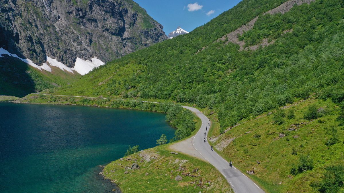 A scenic view of a winding road along a vibrant green hillside and a clear blue lake, bordered by steep mountains with patches of snow. Cyclists traverse the road, enjoying the beautiful landscape under a bright blue sky.