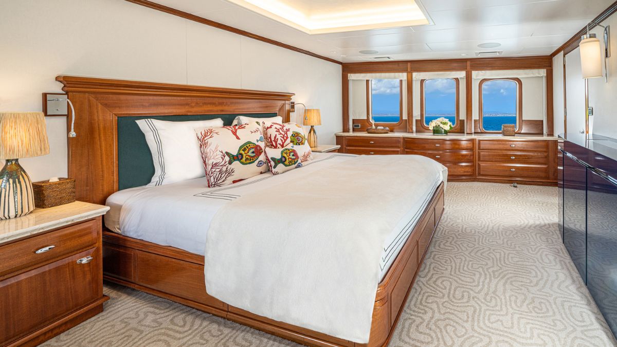A luxurious bedroom featuring a large wooden bed with decorative pillows, bedside lamps, and ocean-view windows. The room has elegant wooden furnishings and a plush rug, creating a serene atmosphere.