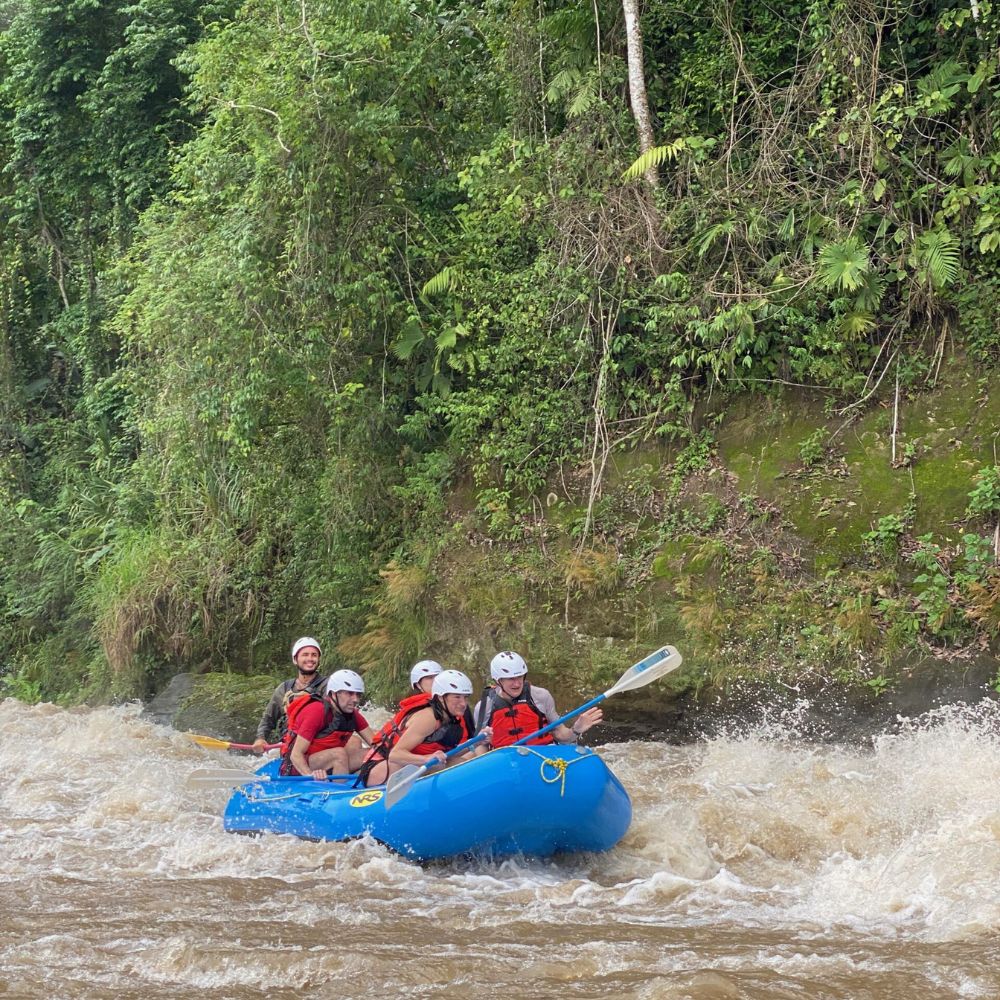 A group of five people wearing helmets and life jackets navigates a blue raft through turbulent waters surrounded by lush greenery.