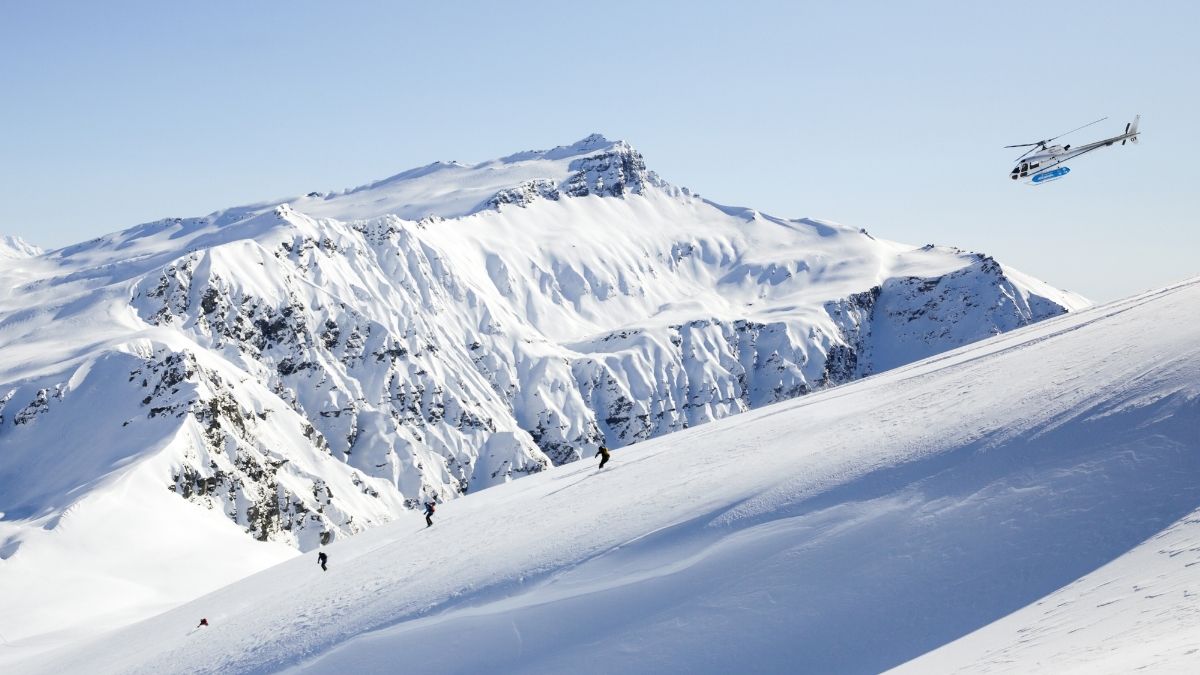 Skiers ascend a snowy mountain slope while a helicopter hovers above against a clear blue sky. Snow-covered peaks rise majestically in the background.