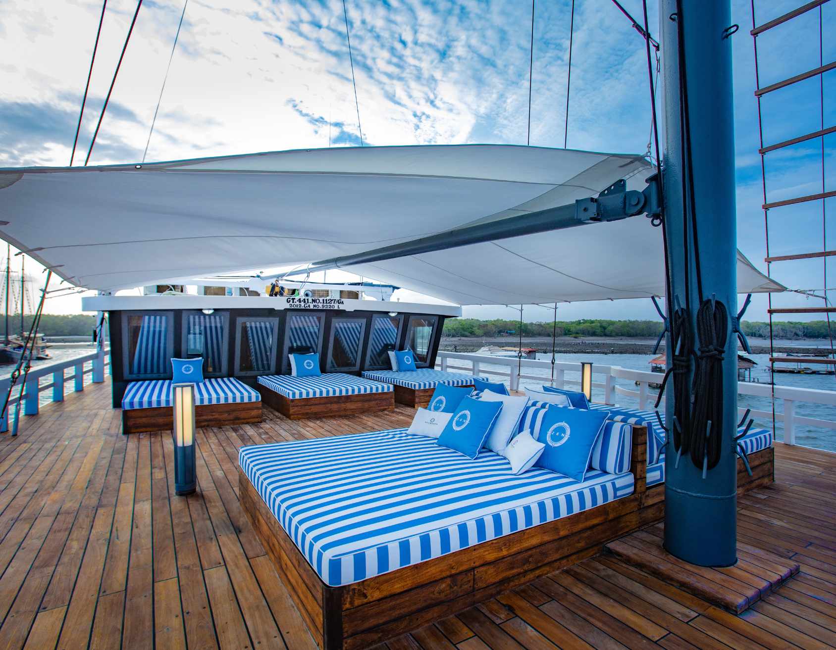Lounge area on a yacht featuring striped cushions and pillows on wooden platforms, shaded by a large canopy. The sky is partly cloudy, creating a serene atmosphere.