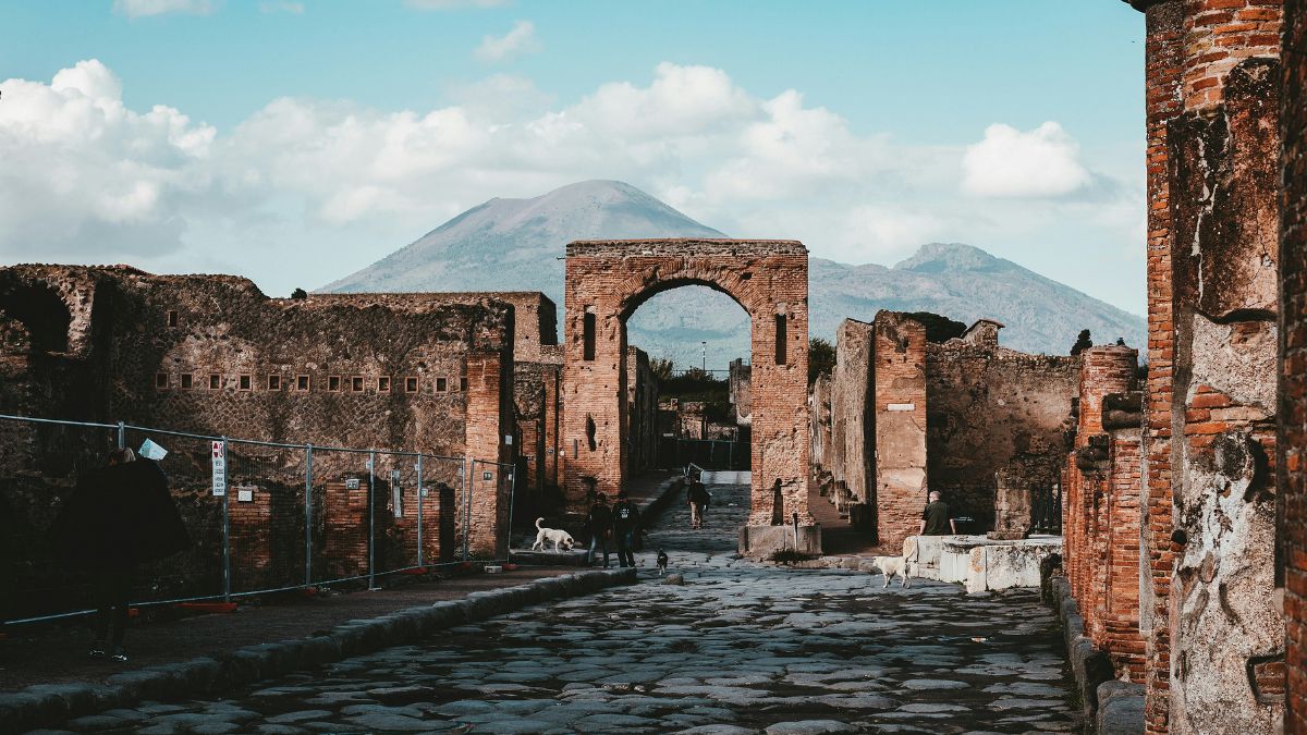 An ancient stone pathway leads through ruins with an archway, framed by weathered brick walls, under a blue sky with fluffy clouds. The silhouette of Mount Vesuvius rises in the background.