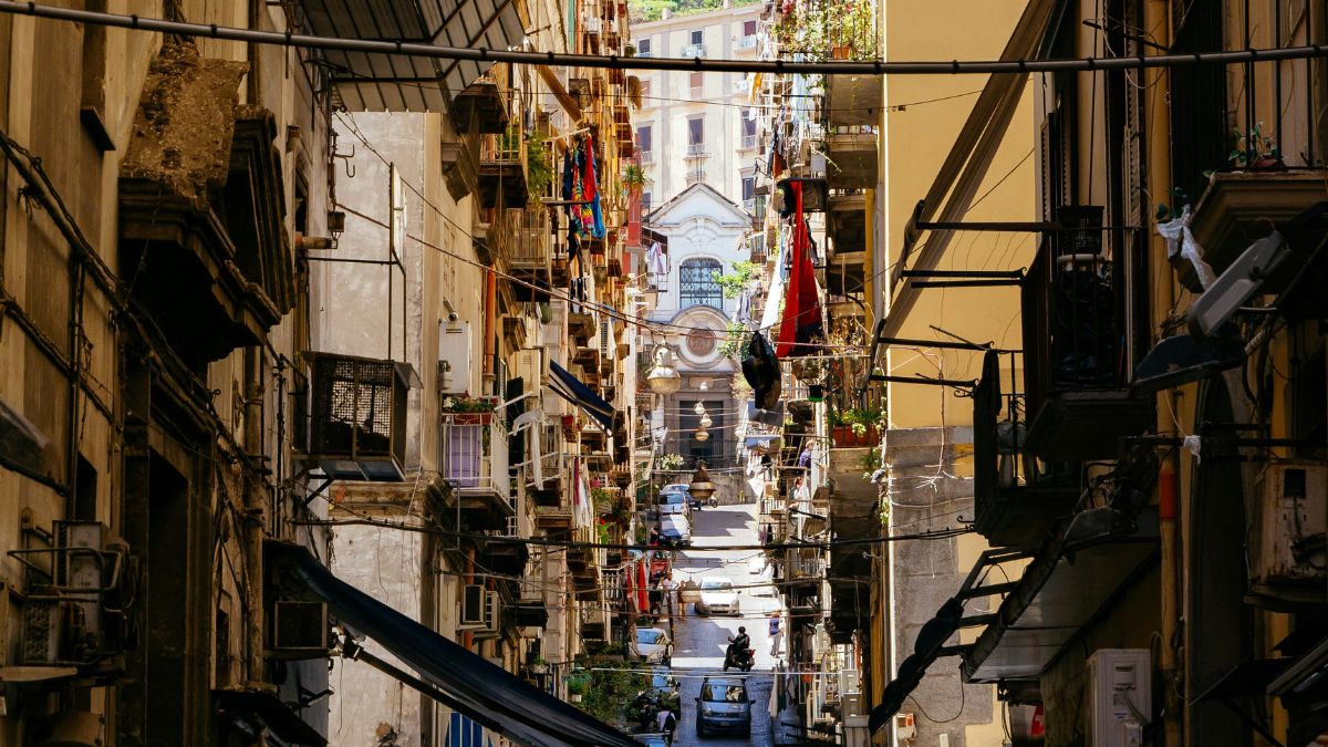 Narrow street lined with colourful buildings and balconies, leading to an illuminated church at the far end. Clothes hang from lines strung across the alley, and a few people can be seen walking.
