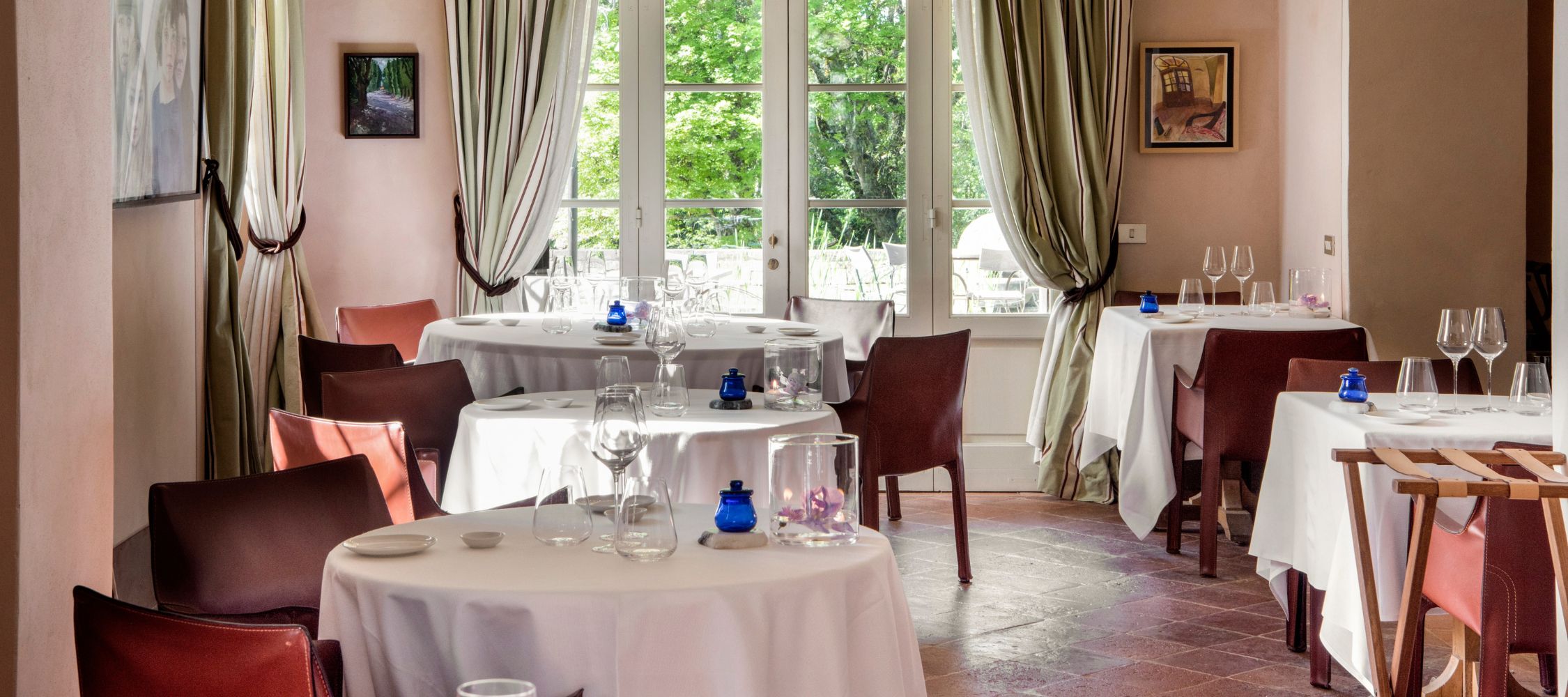 Elegant dining area featuring round tables with white tablecloths, surrounded by wooden chairs. Large windows provide a view of greenery outside, and the space is adorned with soft curtains and tasteful artwork. Wine glasses and decorative elements are arranged on the tables.