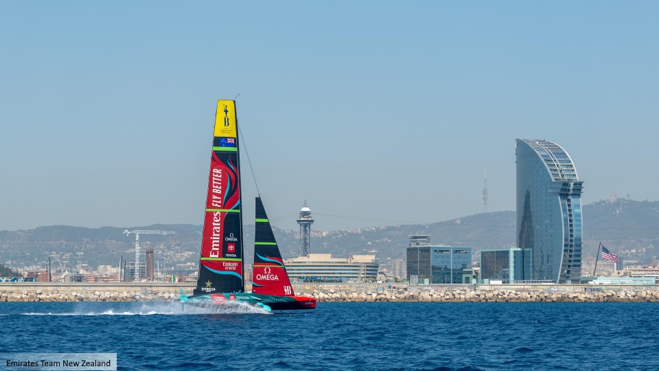 A sleek racing yacht with colorful sails glides through the water near an urban waterfront, featuring modern buildings in the background, including a futuristic tower.