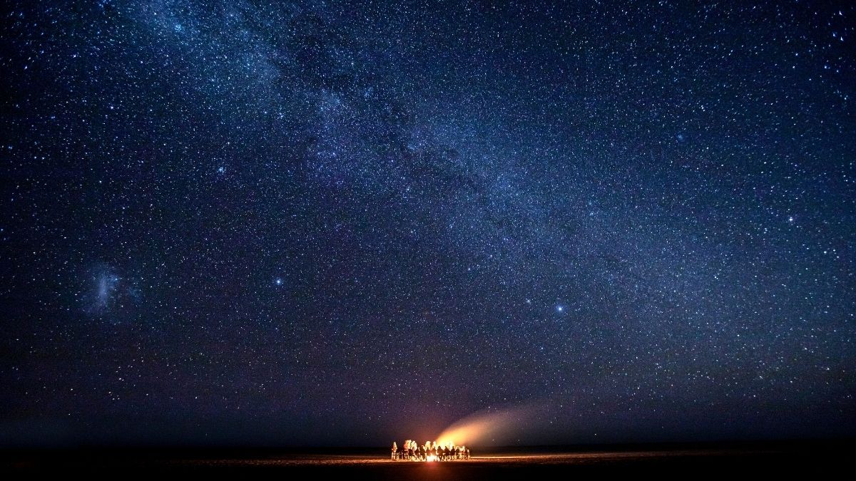 A group of people gathered around a campfire under a starry night sky, with the Milky Way prominently visible above.