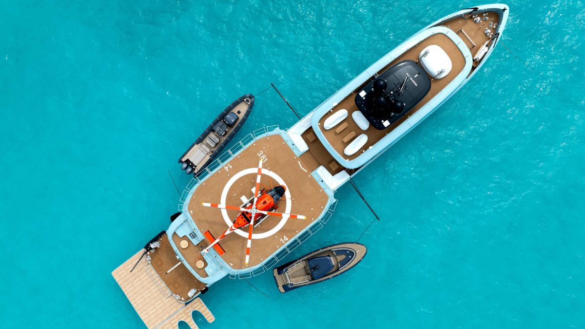 An aerial view of a large boat in clear blue waters with two smaller boats next to it and a red helicopter on the helipad.