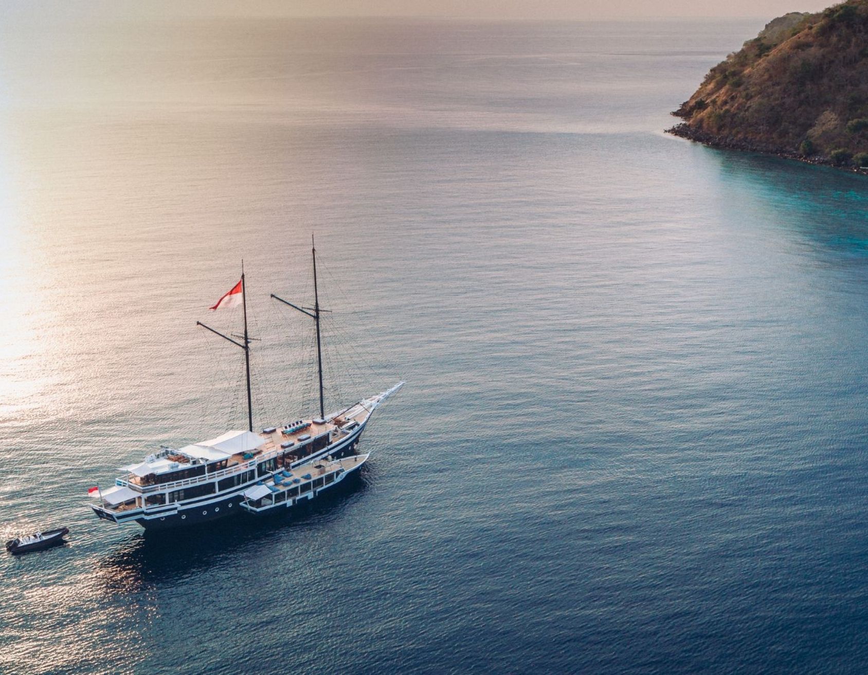 A large sailing ship with multiple masts anchored in calm waters near a rocky coastline. The early morning sky casts a soft glow over the scene.