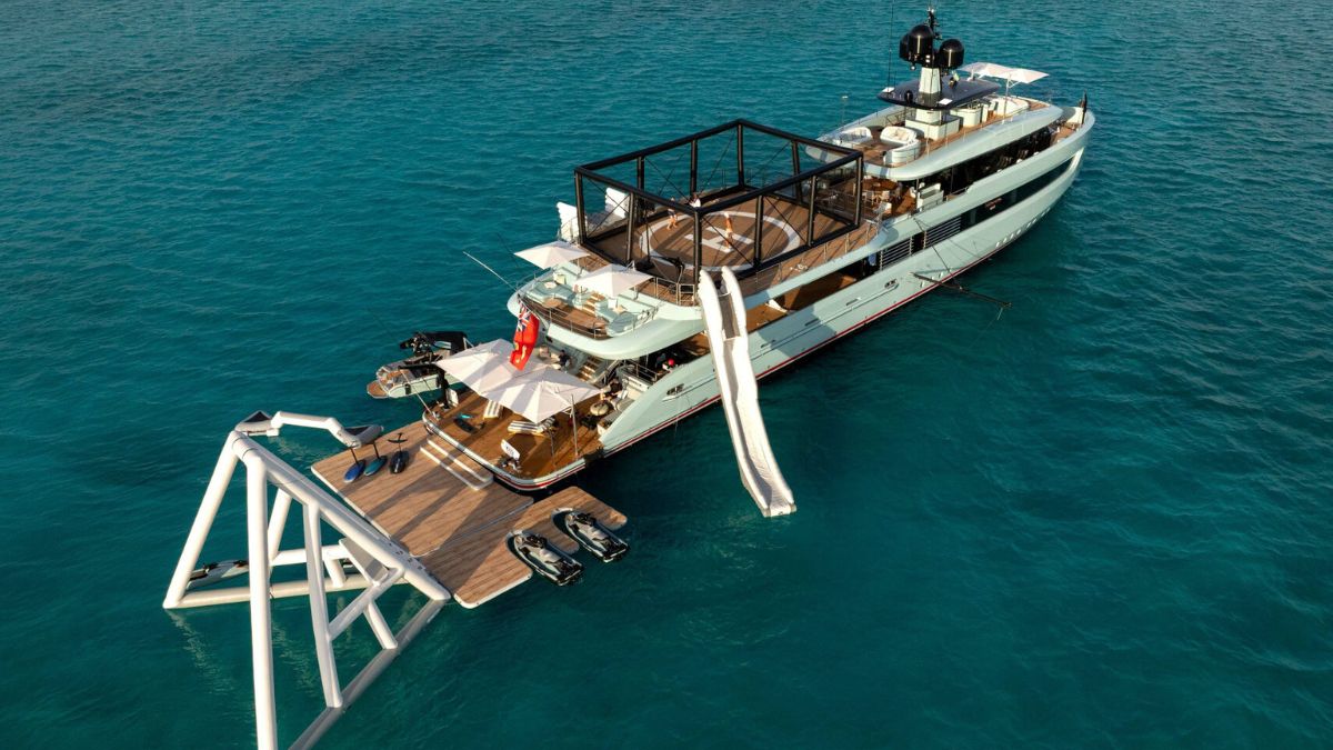 A blue hull luxury yacht in calm blue waters with a sports court, inflatables and slide attached.