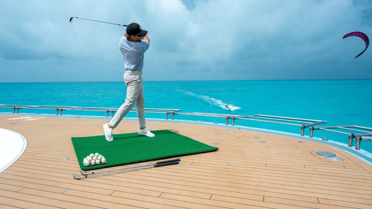 A man on a luxury boat playing golf from a chipping green with golf balls and golf clubs with a clear blue water and kite surfing in the background.