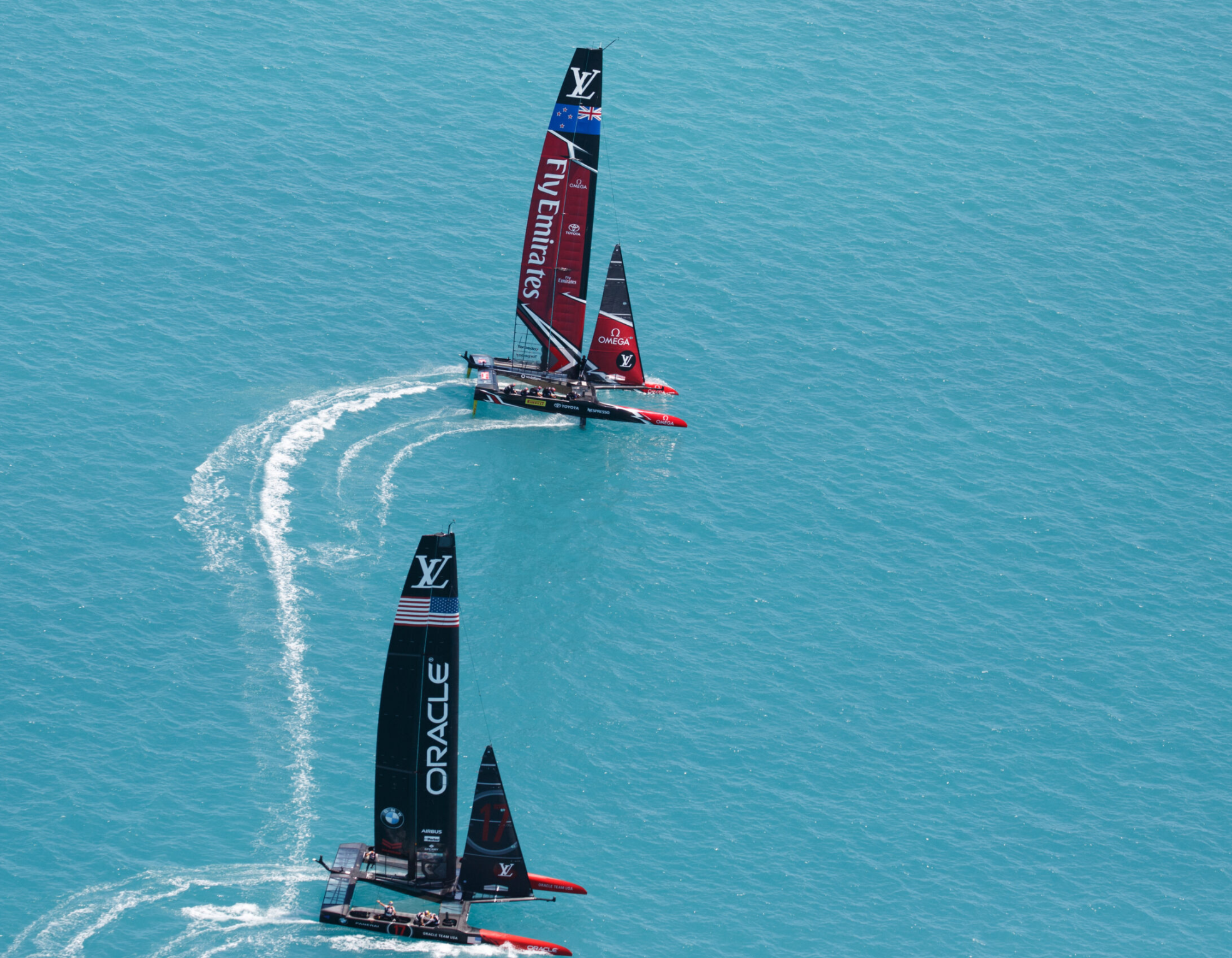 Two racing catamarans glide across vibrant turquoise waters. One is adorned with Emirates Team New Zealand branding, while the other features the Oracle Team logo. The boats leave dynamic white wakes in their path.