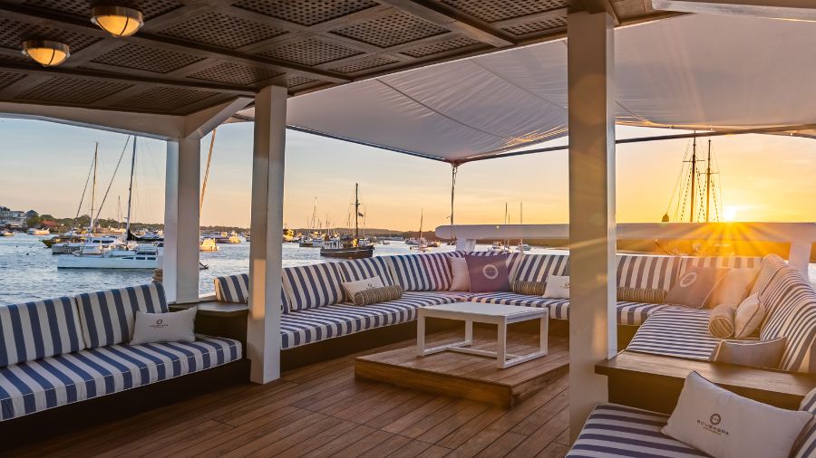A stylish outdoor lounge with striped cushions, overlooking a marina filled with boats. The sun sets on the horizon, casting a warm glow over the scene.