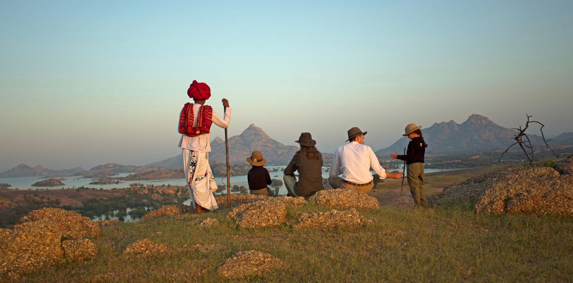 A group of five people, including a man in traditional attire with a red turban and a stick, are standing on a rocky hillside overlooking a scenic landscape of hills and water during sunset. The individuals are dressed in hats and casual clothing, enjoying the view together.