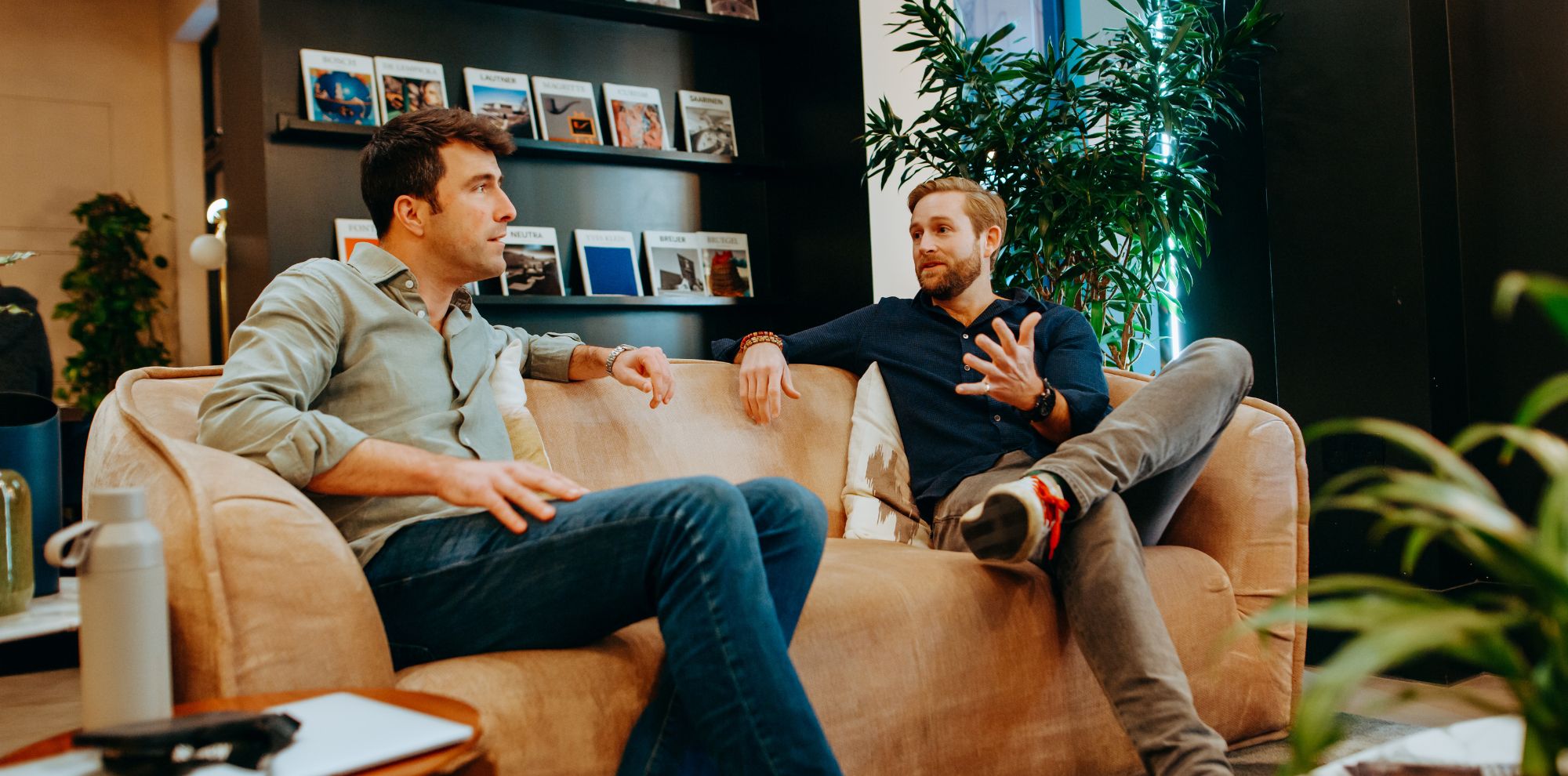 Two men engaged in a conversation while seated on a plush sofa in a modern interior. The background features shelves filled with books and magazines, alongside indoor plants. One man is casually dressed in a light green shirt, while the other wears a dark blue sweater.