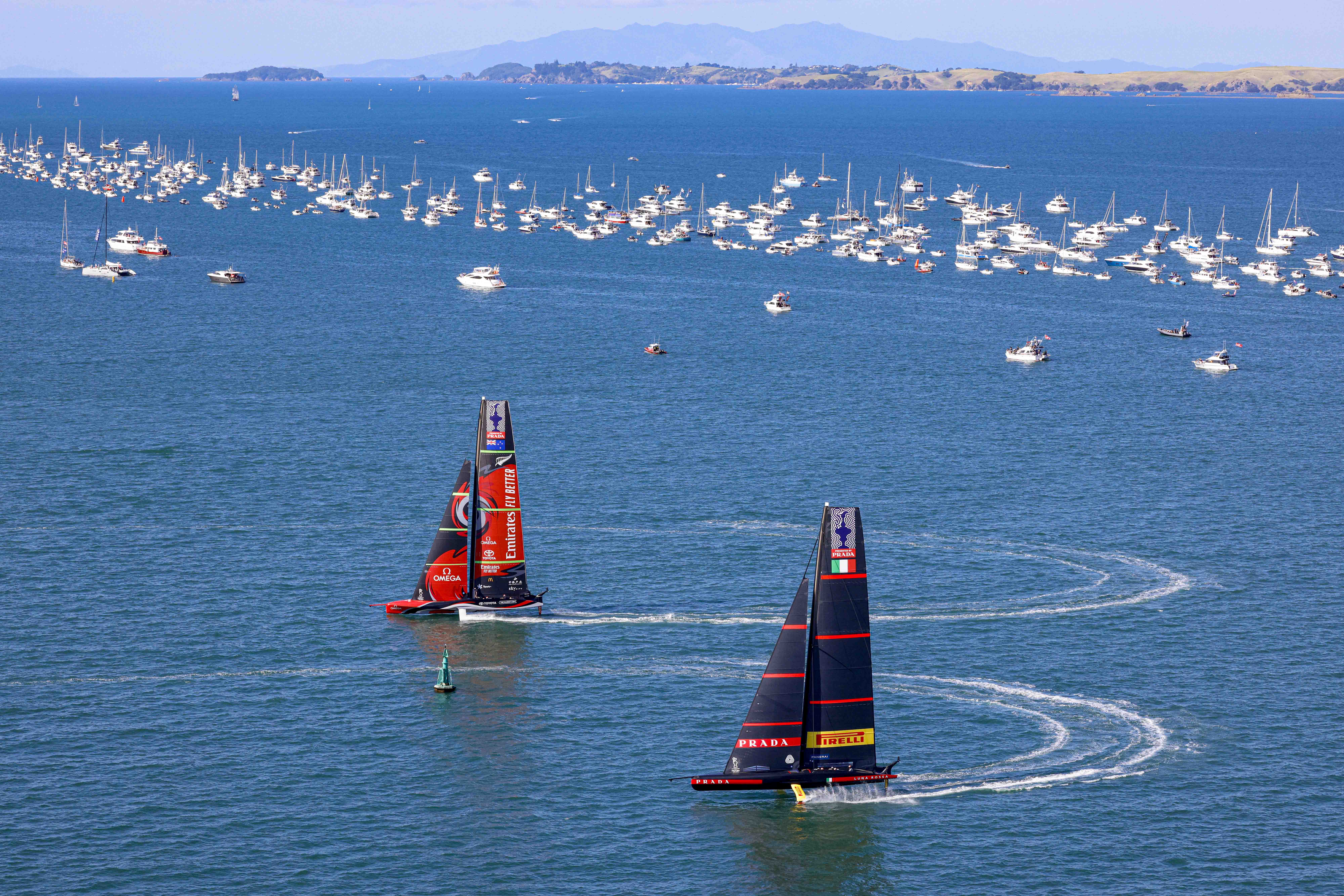 Two racing sailboats navigate the blue waters, while numerous anchored boats are visible in the background. Rolling hills are faintly seen on the horizon under a clear sky.