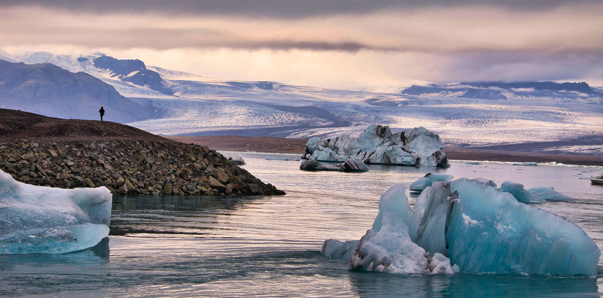 A solitary figure stands on a rocky shoreline, gazing at large icebergs floating in a tranquil blue glacial lagoon. Majestic snow-capped mountains rise in the background under a dramatic sky.