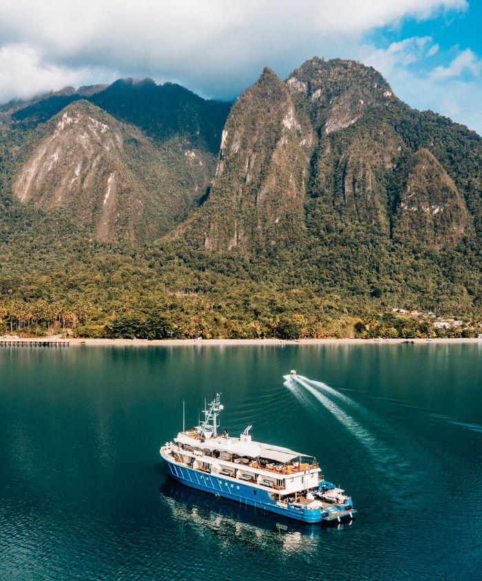 A blue and white yacht sails on calm waters surrounded by lush green mountains under a partly cloudy sky.