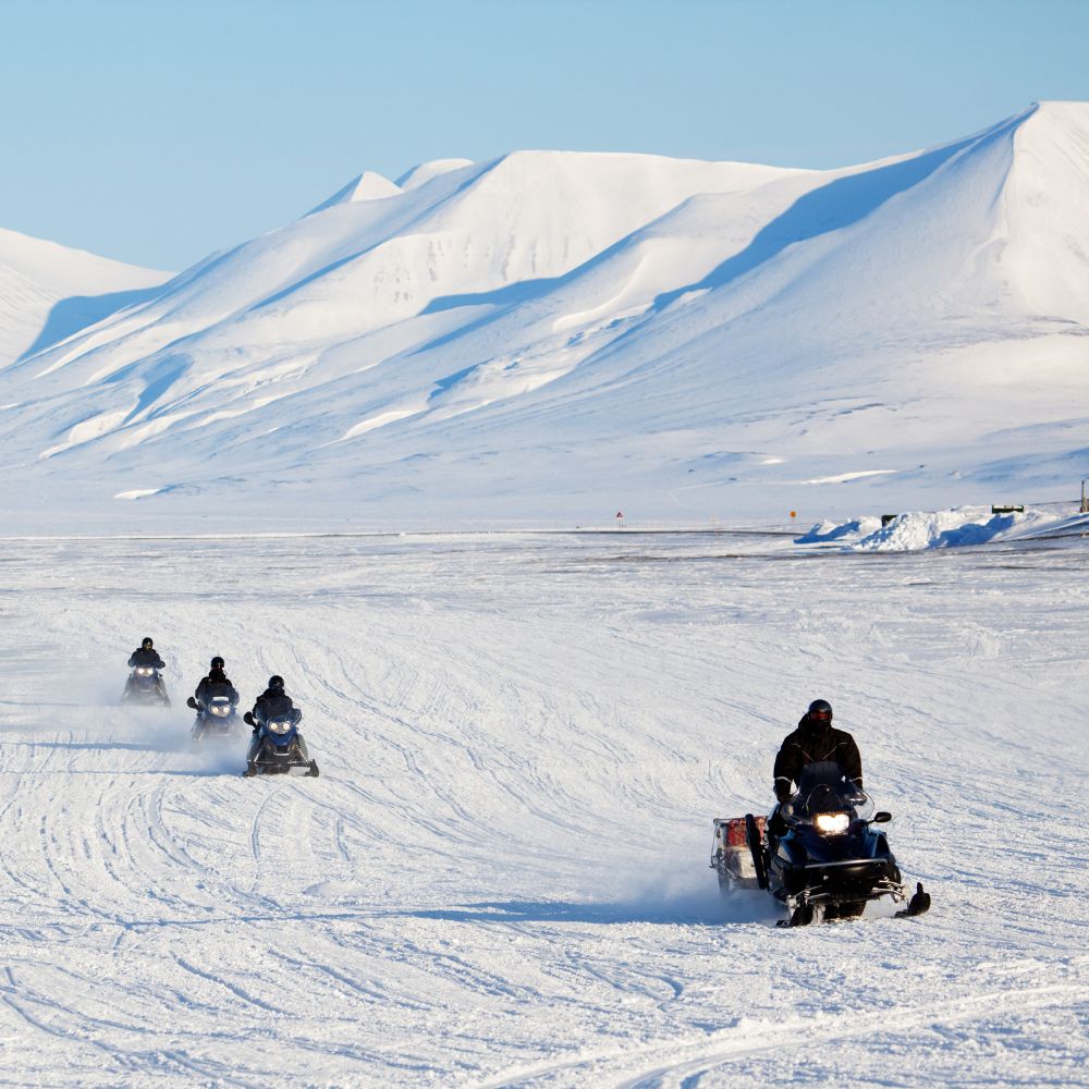 A group of snowmobiles rides across a snowy landscape, with towering white mountains in the background under a clear blue sky.