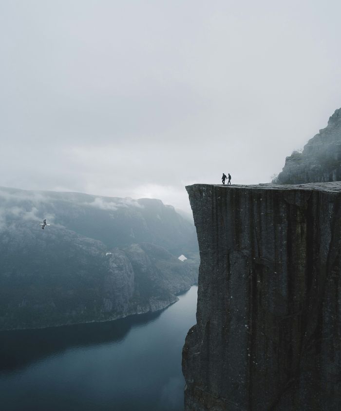 Two figures stand on the edge of a steep cliff overlooking a misty fjord, with mountains and clouds surrounding the serene landscape. A person is seen paragliding in the distance.