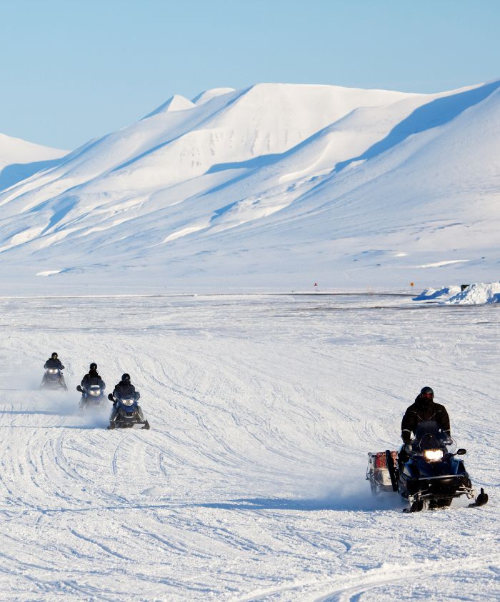 A group of four snowmobiles travels across a snowy landscape with towering white mountains in the background. The clear blue sky contrasts with the bright snow, and trails from the snowmobiles are visible on the surface.