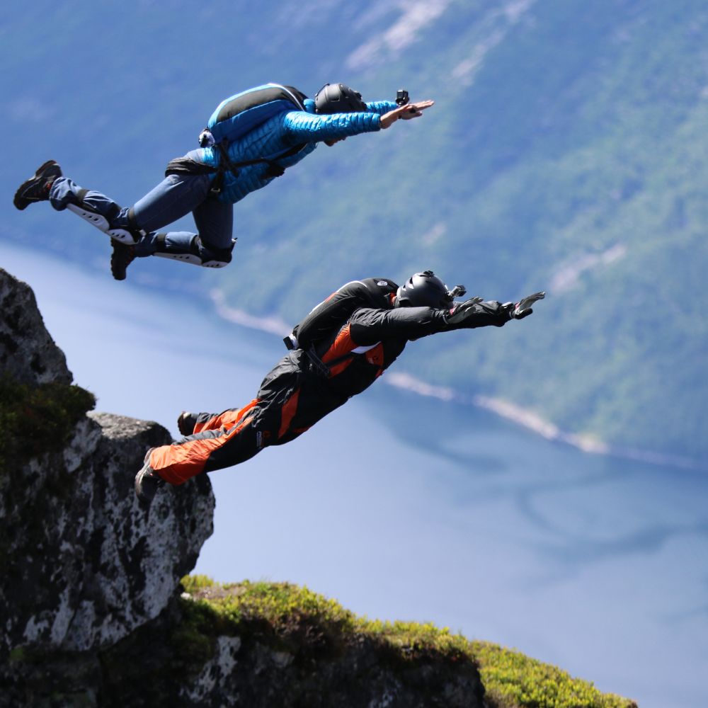 Two base jumpers wearing helmets and jumpsuits leap off a rocky cliff into the air over a scenic fjord. The surrounding landscape features mountains and lush greenery.