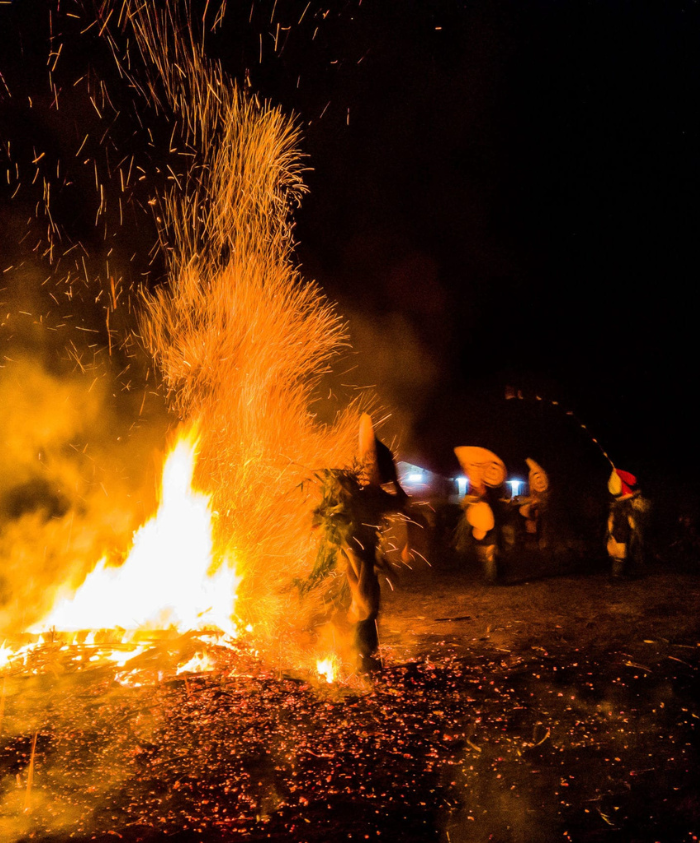 A vibrant fire lights up the night, sending sparks into the air as figures in traditional costumes dance around the flames. The scene is filled with smoke and energy, showcasing a lively celebration.