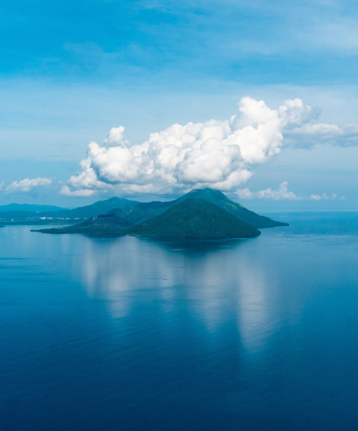 A serene island with a lush green peak rises from calm blue waters under a sky filled with fluffy white clouds.