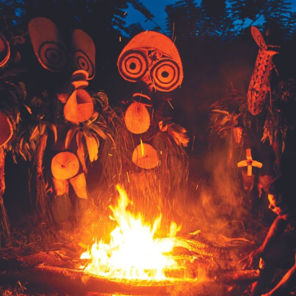 Ceremonial figures made of leaves and woven materials surround a bright fire, creating a mystical atmosphere at dusk. The figures have large, expressive faces and are adorned with various natural elements.