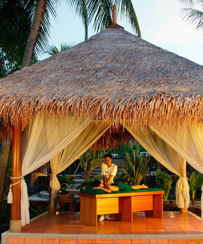 A serene outdoor spa gazebo with a thatched roof, featuring a massage table and a therapist preparing for a treatment amidst lush tropical greenery.