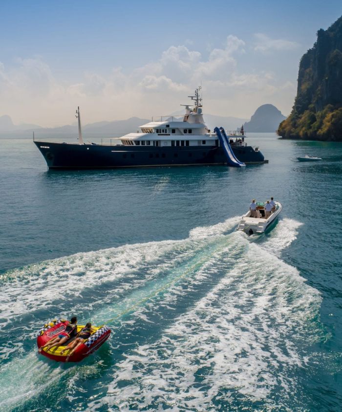 A large luxury yacht anchored in calm waters with a waterslide attached, while a small motorboat tows two people on a banana boat in the foreground, surrounded by rugged cliffs and a clear blue sky.