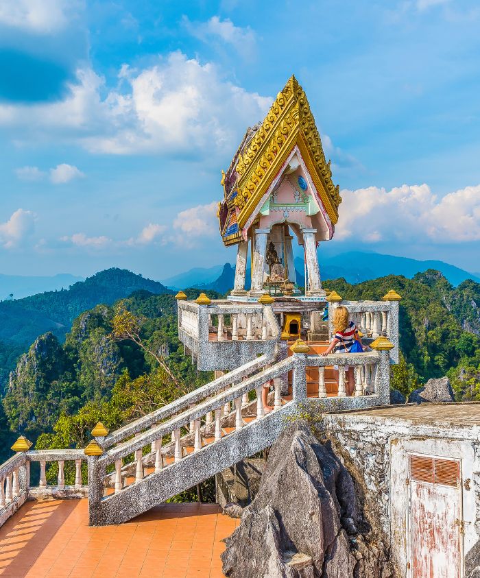 A small temple with a colorful roof sits atop a rocky peak, surrounded by lush mountains and a blue sky. A person rests on the stairs leading to the temple.