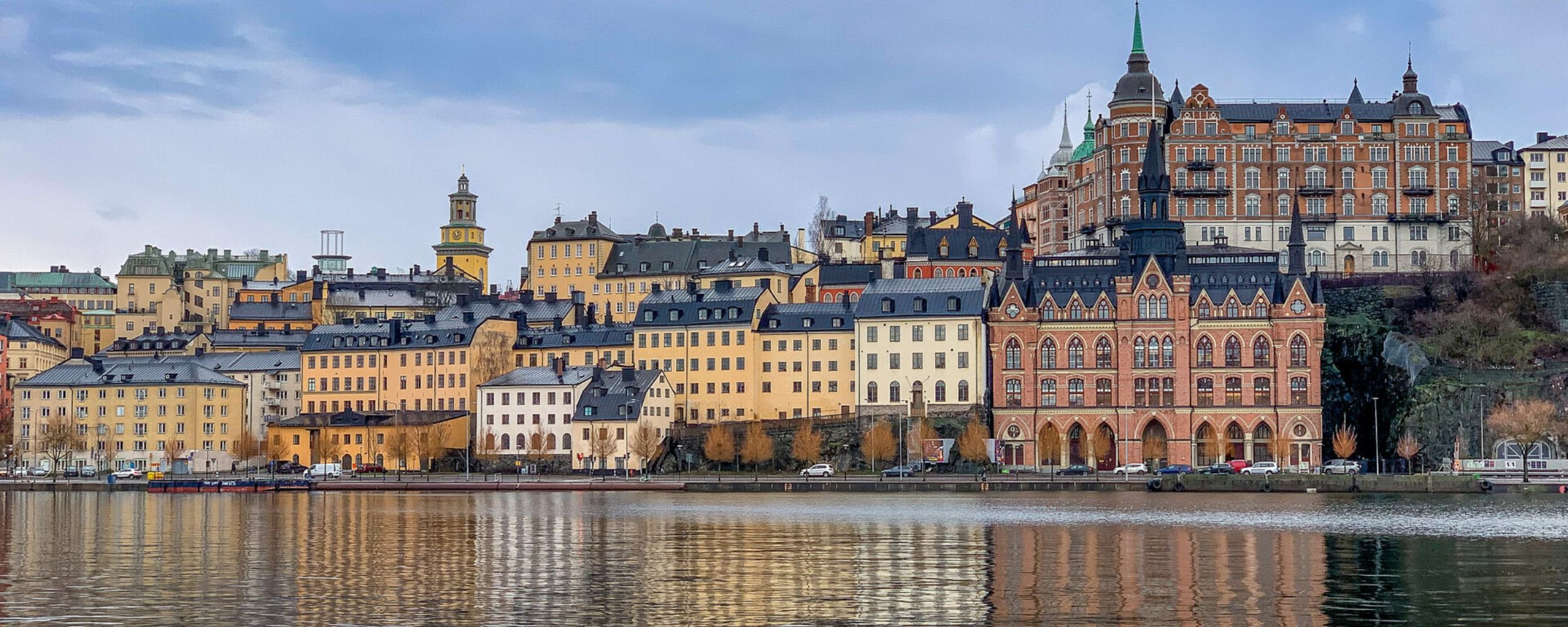 A scenic view of a waterfront in Stockholm, showcasing colorful historic buildings along the shore, with reflections in the calm water. Prominent structures include a mix of architectural styles, with spires and detailed facades, under a cloudy sky.