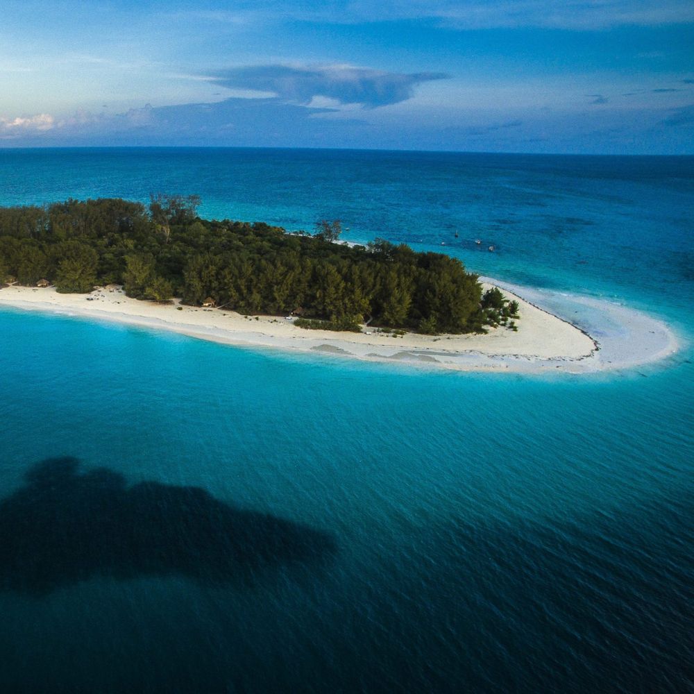 Aerial view of a small, tropical island surrounded by clear turquoise waters and a sandy beach. Lush greenery covers the island, with a gentle curve of white sand contrasting the vibrant sea.