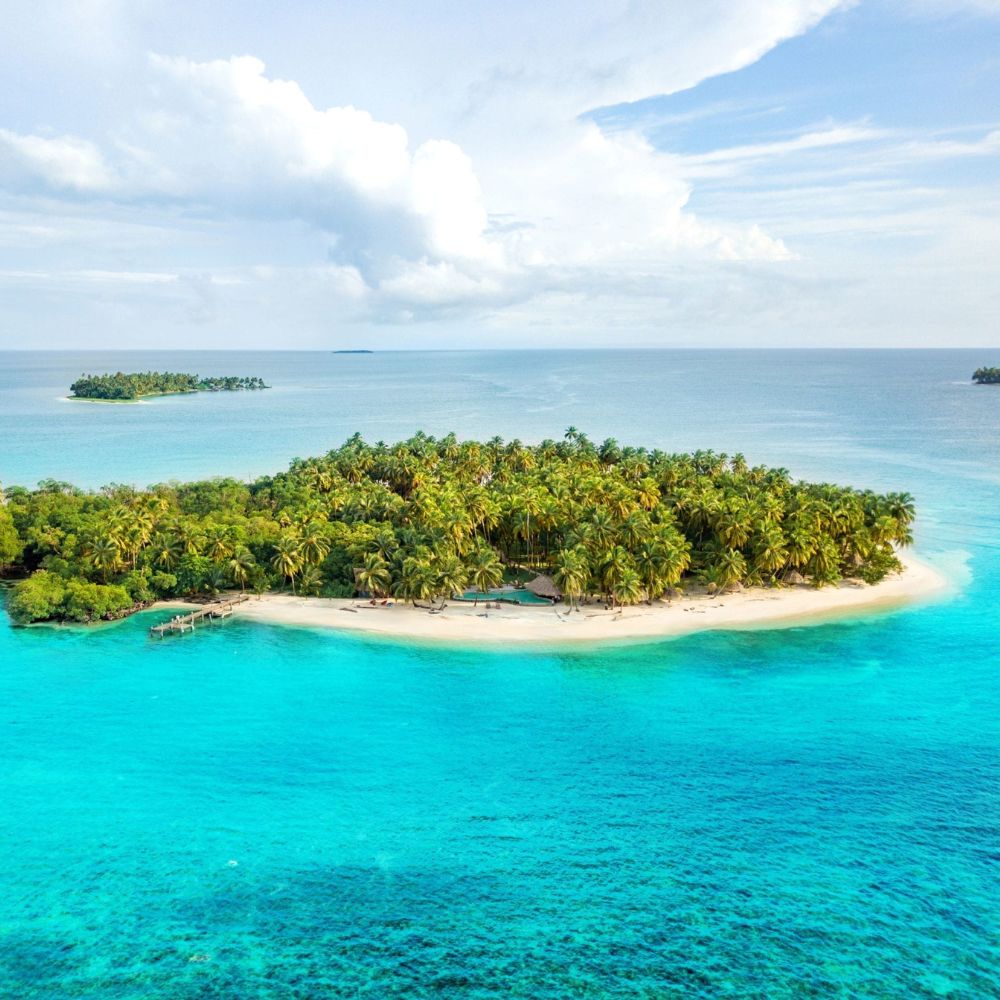 Aerial view of a tropical island surrounded by turquoise waters, featuring lush palm trees and a sandy beach, with smaller islands visible in the distance.