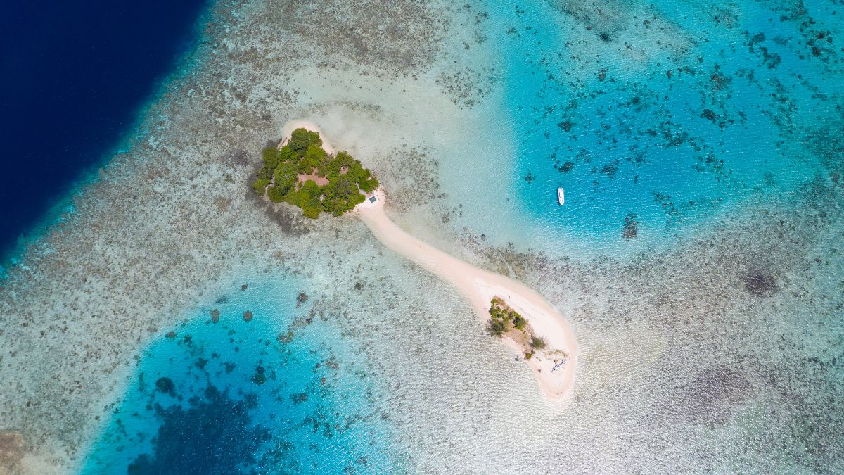 Aerial view of a small, sandy island connected by a narrow strip of beach, surrounded by vibrant turquoise waters and coral reefs. A small boat is visible near the island.