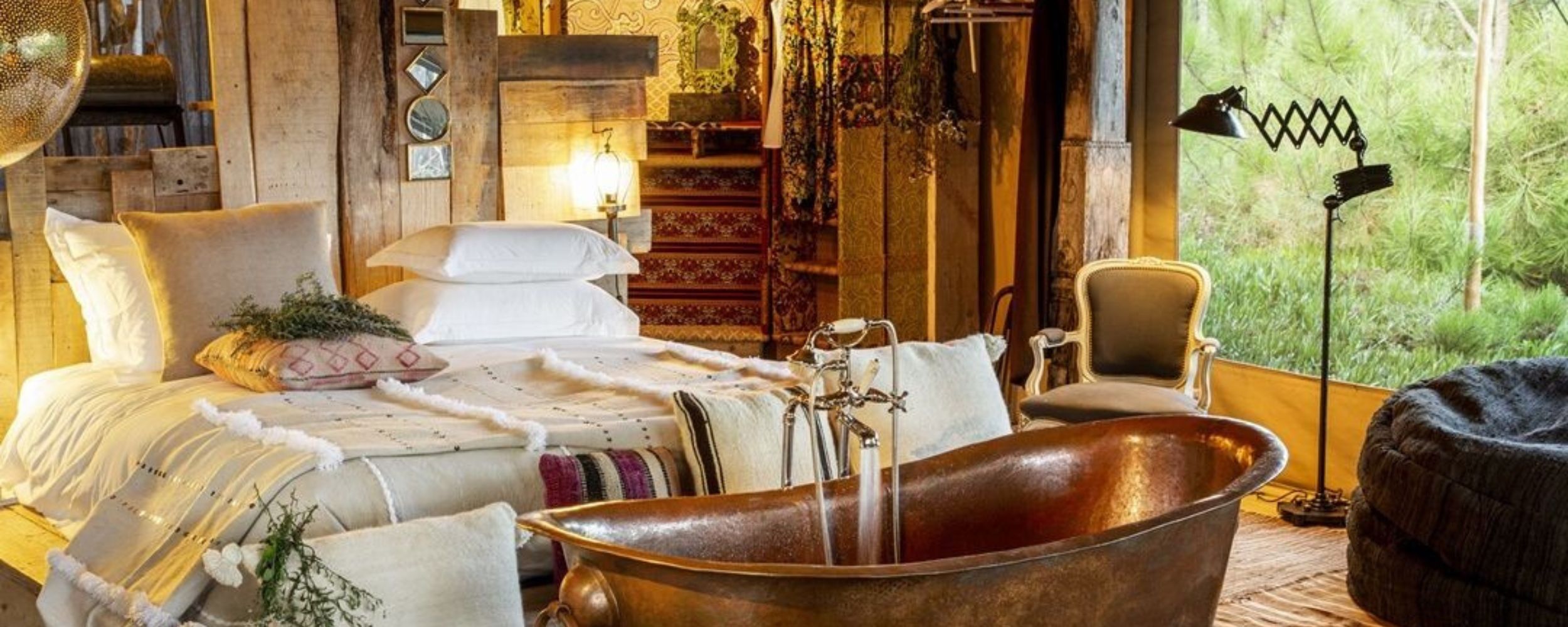 A cozy, rustic bedroom featuring a large copper bathtub, a comfortable bed with plush pillows, and a vintage chair. Natural light floods in from large windows showcasing greenery outside. Decor includes textured fabrics and decorative accents, creating a warm and inviting atmosphere.