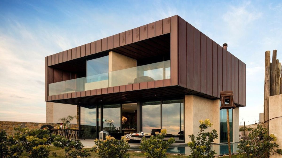 A modern two-story house with a sleek metal exterior and large glass windows. The building features a balcony on the upper floor and is surrounded by greenery. A swimming pool is visible in front, reflecting the clear sky.
