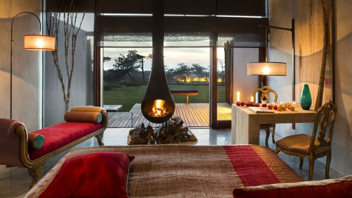 Cozy bedroom with a modern fireplace, featuring a red bedspread and accent pillows. A lounge area with a red bench and a wooden table decorated with fruits. Large windows offer a view of an outdoor space with greenery. Warm ambient lighting creates a welcoming atmosphere.