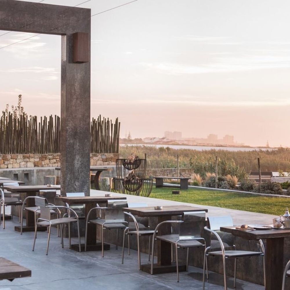 An open-air dining area features modern metallic tables and chairs, surrounded by greenery and sculptures, with a sunset view over the horizon.