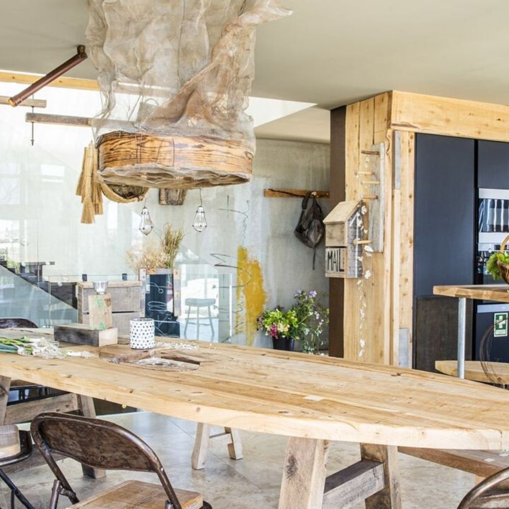 A modern wooden dining table with rustic chairs in a contemporary kitchen, featuring a unique hanging light fixture made of natural materials. The space has glass walls, decorative plants, and a minimalist design.