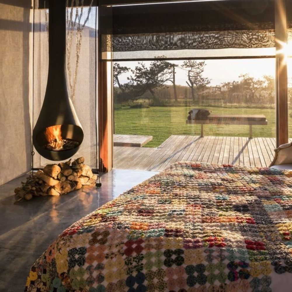 A cozy bedroom featuring a colorful quilt on the bed, with a modern fireplace hanging from the ceiling. Large windows showcase a serene outdoor view and natural light streaming in.